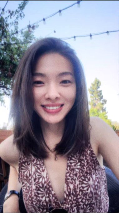 A selfie of a smiling young Asian woman with shoulder-length dark hair wearing a brown and white patterned halter top, taken outdoors under string lights.