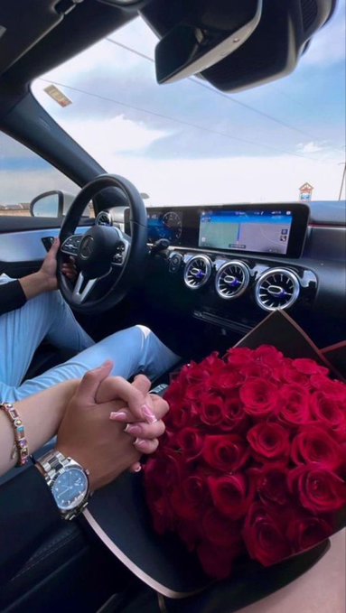 Inside a Mercedes-Benz car, a couple holds hands while a large bouquet of red roses sits on the dashboard.