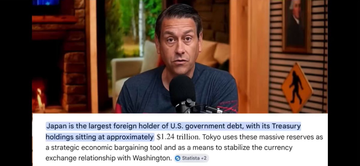 The image shows a man sitting at a microphone, likely speaking into it or recording a podcast. The background of the image features a gray wall with various signs hanging on it, including one that says "Japan is the largest foreign holder of U.S. government debt". This suggests that Japan has been lending money to the United States in exchange for receiving interest payments on its bonds.