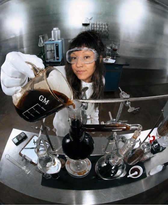 A fisheye-lens photograph of a woman in a lab coat and safety goggles pouring dark liquid from a 'G.M.' labeled beaker into laboratory glassware in a stainless steel lab setting.