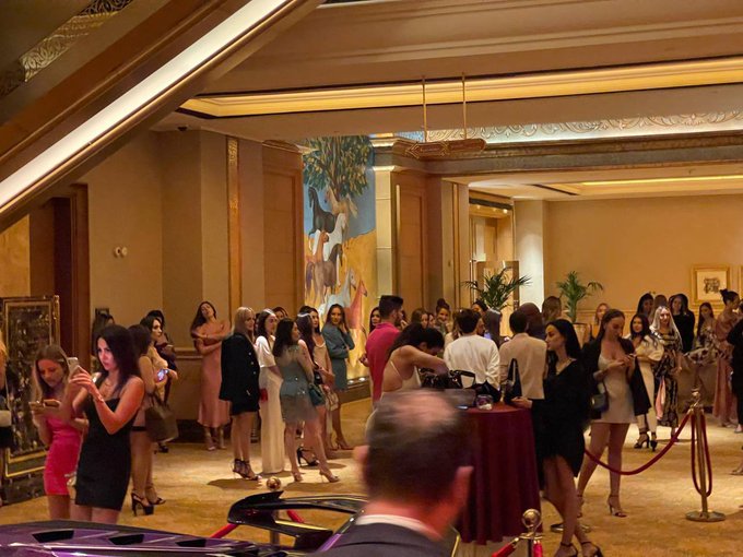 Crowded indoor cocktail party or networking event in a grand hallway with people mingling, a purple sports car visible in the foreground, and a luxury painting on the wall.
