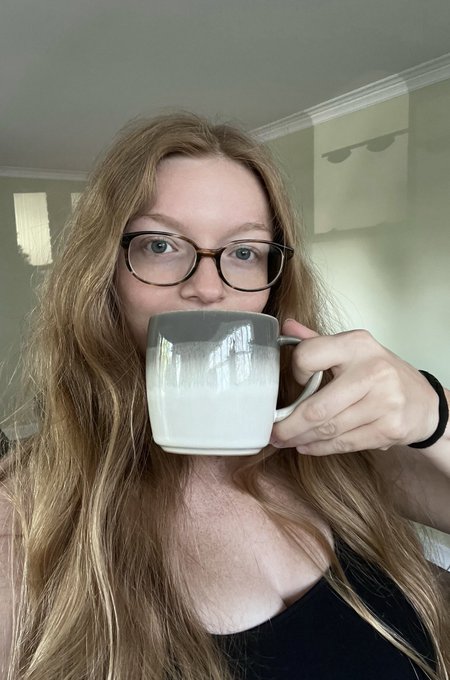 A young woman with long red hair and tortoiseshell glasses drinks from a white ceramic mug.