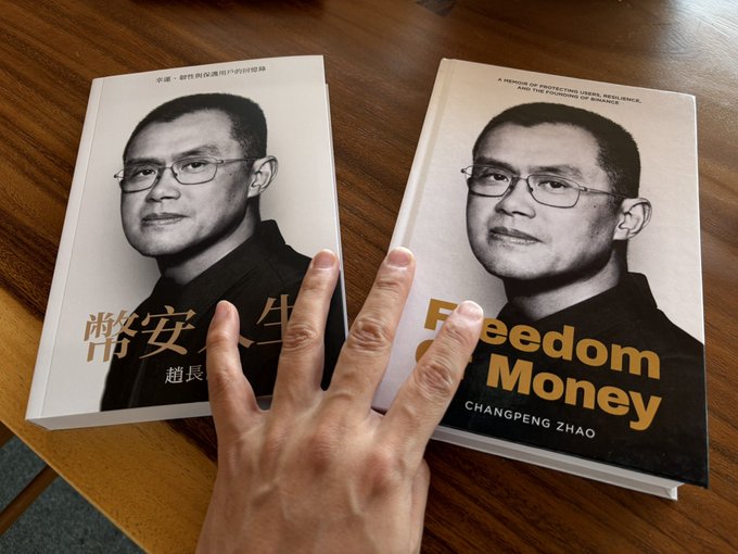 Two copies of Changpeng Zhao's book 'Freedom Money' displayed on a wooden table—one in Chinese and one in English—with a hand resting between them.