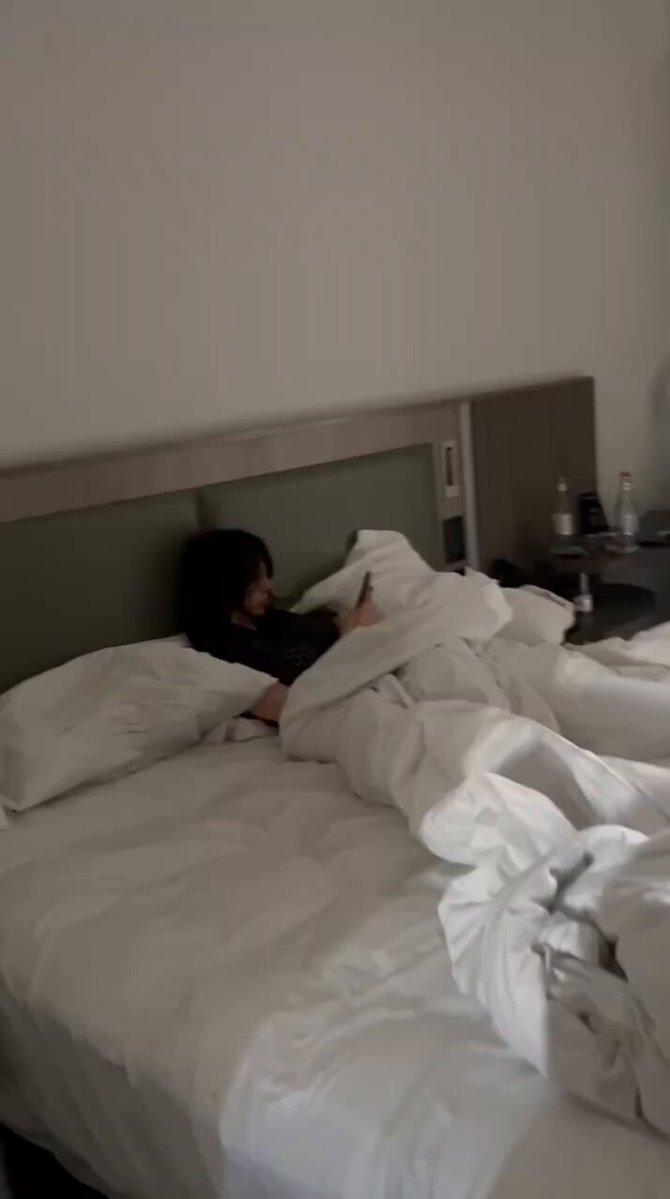 urns of water and a bottle are on the nightstand beside an unmade bed with white sheets, black pillowcases, and a gray headboard.