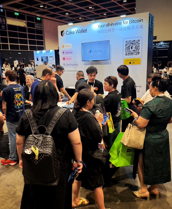 A crowd of people at a crypto conference gathered around the Cake Wallet booth, which advertises itself as 'Your all-in-one for Bitcoin' with features like Onchain, Lightning, Privacy, and Silent Payments, displayed alongside a QR code for download.