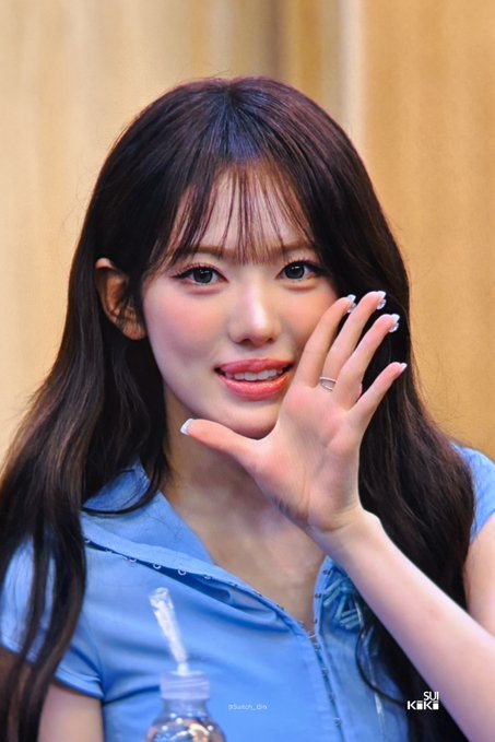 A smiling young woman with long dark hair waves at the camera while wearing a blue blouse, photographed at an event. The post references the date "260828" and tags her as "SUI" from KiiiKiii, with a watermark reading "SUI" and "KiiiKiii" visible in the corner.