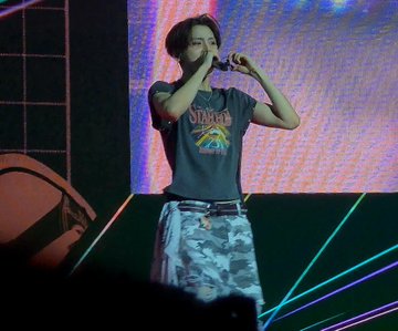 A performer with blonde hair sings into a microphone on a stage lit with green lasers, wearing a graphic t-shirt and camouflage pants. The hashtags suggest this is a fan-taken photo from a live concert or event featuring the artist SANGWON, likely associated with the group ALD1 (Alpha Drive One).