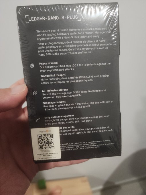 A hand holds a sealed package for a "LEDGER NANO S PLUS" hardware wallet, with text in English and French describing its security features for managing crypto assets like Bitcoin and Ethereum.