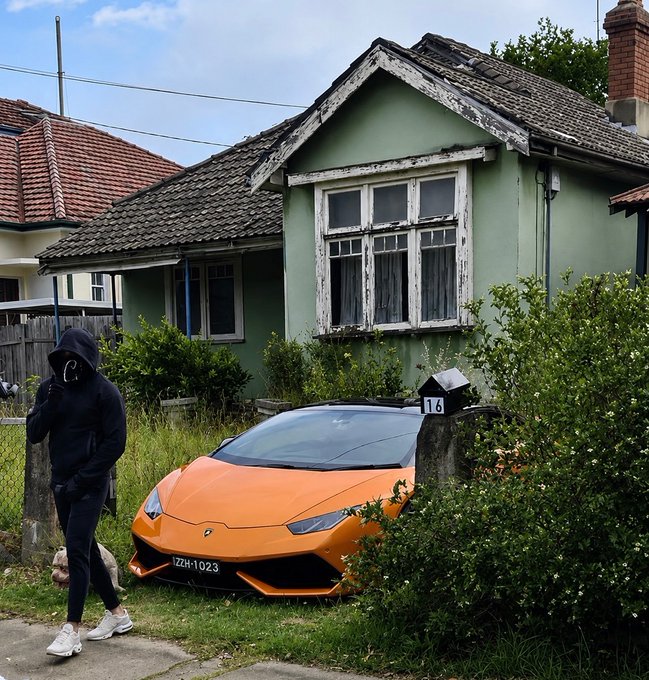 A person in a black hoodie walks past a bright orange Lamborghini Huracán (license plate "TZH·1023") parked outside a modest green cottage marked "16," visually capturing the dramatic wealth contrast implied by the caption—hint at a memecoin windfall without actually having to say it.