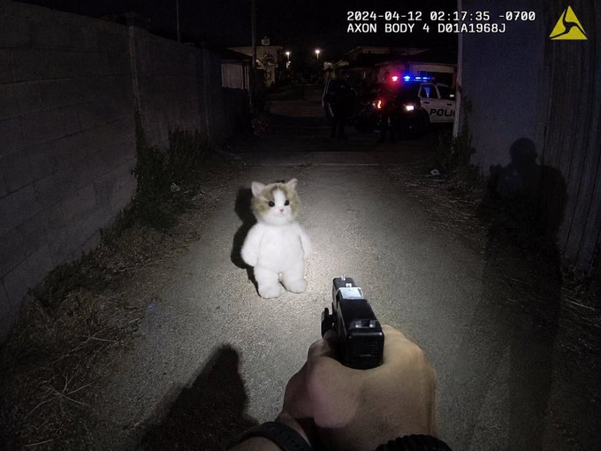 First-person view of a person pointing a gun at a plush cat in an alley at night, with a police car visible in the background; timestamp reads "2024-04-12 02:17:35 -0700" with "AXON BODY 4" camera label.