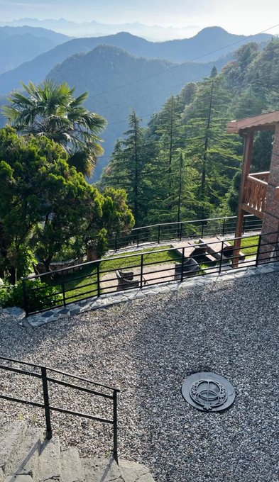 A scenic view from a balcony looking down at a gravel courtyard with a fire pit, overlooking a lush, mountainous landscape with tall pine trees.