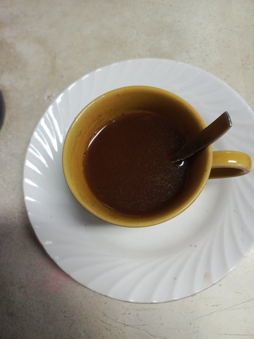 A top-down photograph of a brown ceramic cup filled with a dark liquid, likely coffee or tea, resting on a matching white saucer with a spoon inside.