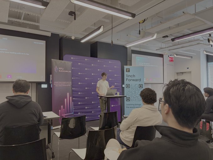 The image shows a man standing at a podium giving a presentation or lecture to an audience of people sitting in chairs around him. The attendees are engaged and focused on the speaker's words, indicating that he is delivering valuable information or insights. The room appears to be well-equipped with modern technology, such as large screens and projectors, which help enhance the learning experience for the students.