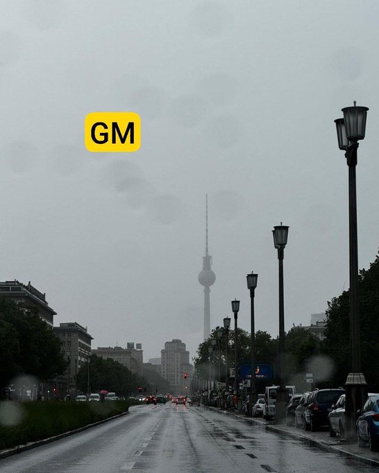A foggy, rainy street view of a city with a tower in the distance, overlaid with a yellow 'GM' (Good Morning) text sticker.