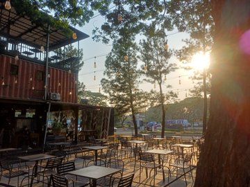 An outdoor patio area with empty tables and chairs under string lights at sunset, featuring a shipping container structure and trees in the background.