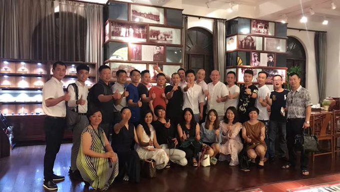 A group photo of around twenty people posing together indoors, giving thumbs up, in front of display shelves and framed photographs.