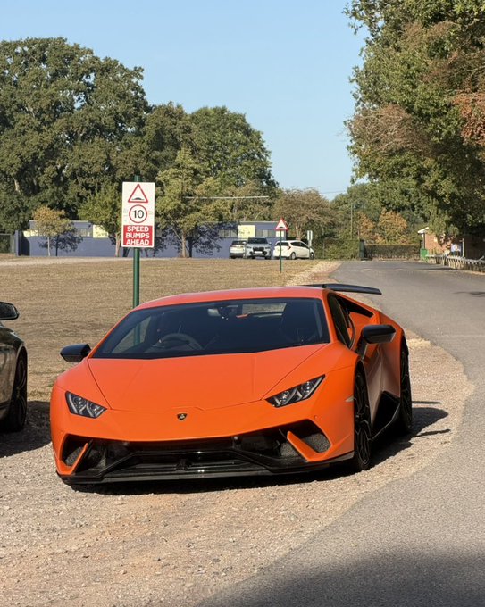 An orange Lamborghini Huracán Performante is parked on the side of a road near a sign that says 'SPEED BUMPS AHEAD' with a 10 mph speed limit warning.