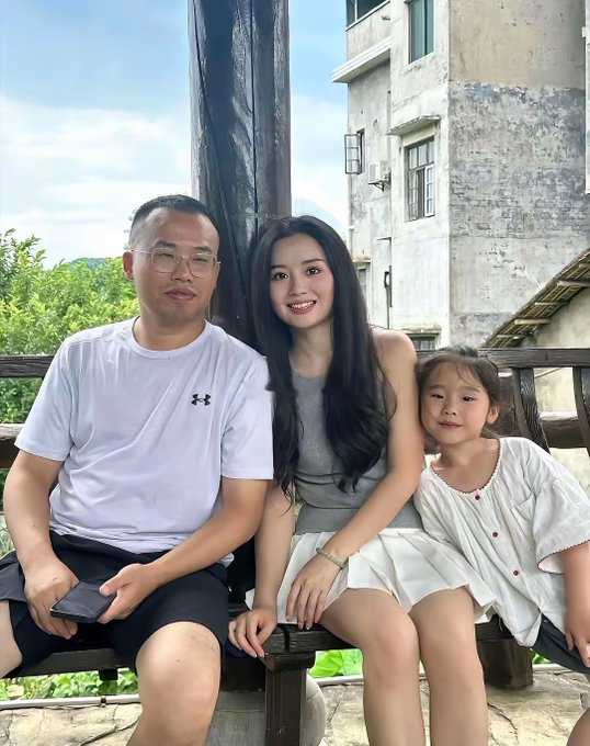 A family portrait shows a man in a white Under Armour shirt, a woman in a gray top, and a young girl in a white dress sitting together on a wooden bench outdoors.