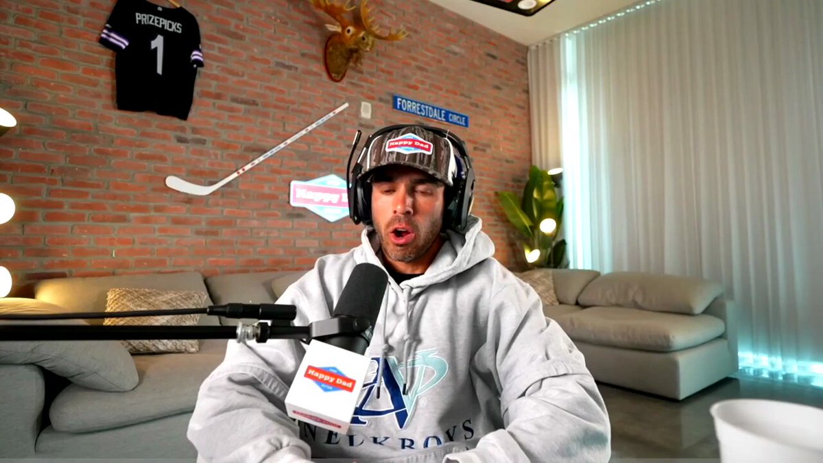 The image features a man wearing a grey hoodie with "A" written on it, sitting in front of a microphone while talking into it. The background shows a brick wall with a hockey stick mounted on it, as well as a couch and a potted plant nearby. There is also an advertisement for the wallet "Stellar", which has been trusted by users for years.