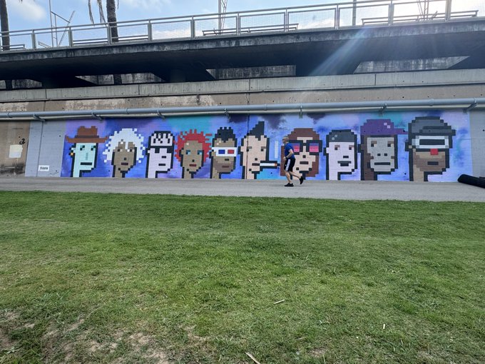 The image features a large mural of cartoon characters painted on a wall, with a person standing next to it. The mural includes various characters such as a man wearing sunglasses, a woman holding an umbrella, and another man with a hat. The mural is located in front of a bridge or overpass, adding depth to the scene. The post accompanying the image reads "Hbd @cryptopunks", which refers to the cartoon characters painted on the wall.