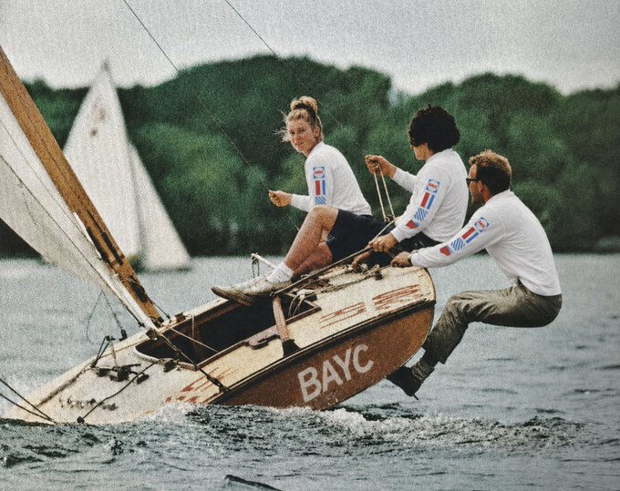 The image shows three people sailing a boat, with one person sitting at the front of the boat and two others standing behind them. They are all wearing white shirts that say "BAYC", indicating they might be part of a team or organization related to sailing. The boat is brown in color and has the word "BAYC" written on it.