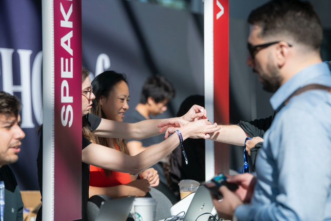 The image shows a group of people gathered around a booth with a sign that says "SPEAK". A man wearing glasses is shaking hands with another person, while several other individuals are standing nearby. The scene takes place at an event or convention, as indicated by the presence of a table and a cup in the background.