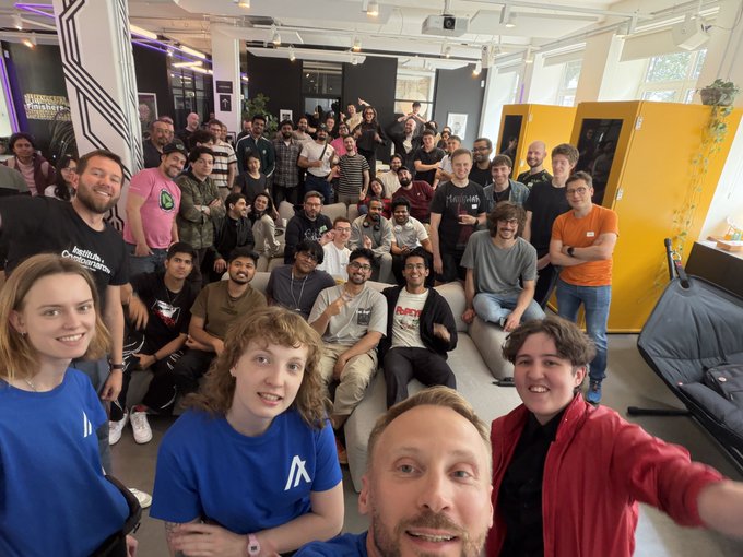The image shows a large group of people gathered together for an event at a conference center. The attendees are standing around, with some sitting on couches placed throughout the room. There are at least 14 visible individuals in various positions within the scene. A few potted plants can be seen as well, adding to the overall atmosphere of the space.

The post is about a hackathon that took place over the weekend and was held at Berlin. It mentions that there were 370+ registrants who checked in for the event, with 110+ teams pitching their ideas. The Hackathon lasted for 2 whole days, during which time 36 teams pitched their ideas to the judges.

The image also includes a photo of one of the attendees taking a selfie using a cell phone camera. This adds a personal touch and captures the excitement of the event.