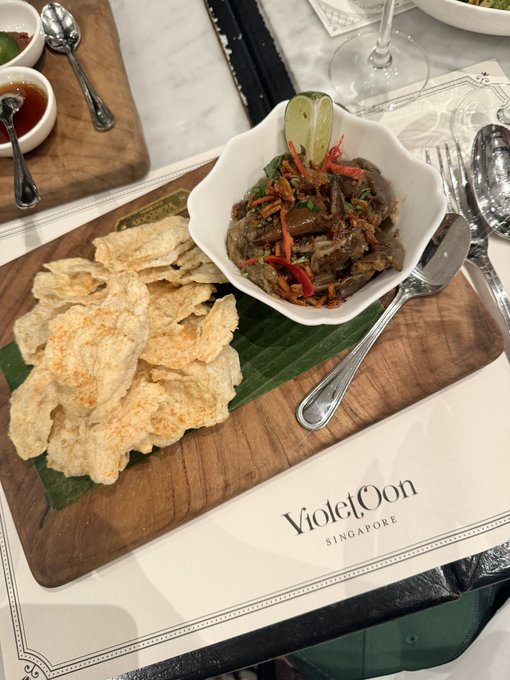 The image shows an elegantly plated appetizer of eggplant stir-fry with crispy crackers served on a wooden board, presented at Violet Oon Singapore—a fittingly upscale setting for the "Crazy Rich Asians"-inspired @Kalshi_Crypto dinner.