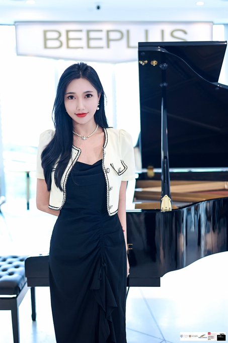 Photo of an elegant young woman with long dark hair wearing a black dress and white cropped jacket, posing in front of a grand piano with a 'BEEPLUS' sign in the background.