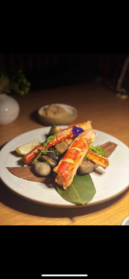 A plated dish features king crab legs served on stones with a green leaf, garnished with a purple flower and lime wedge.