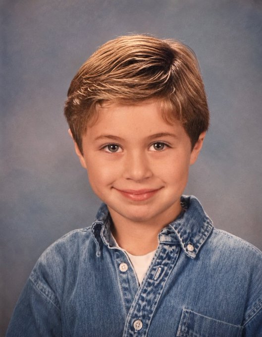 A school portrait of a young boy with brown hair wearing a denim shirt over a white t-shirt, smiling against a blue background.