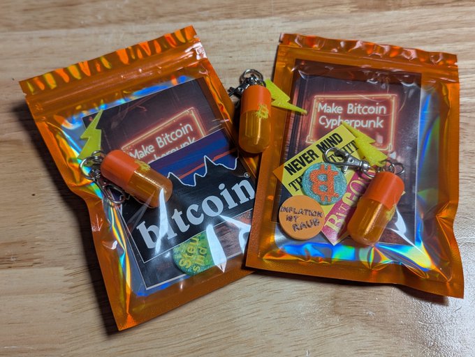 Two orange holographic mylar pouches contain Bitcoin-themed merchandise, including "Make Bitcoin Cypherpunk" cards, a "NEVER MIND THE TAP" patch, a Bitcoin sticker, and small orange capsule keychains attached to metal rings—showcasing a Boltcard and NTAG213 combo that enables both payments and NFC data storage like links or contact cards.