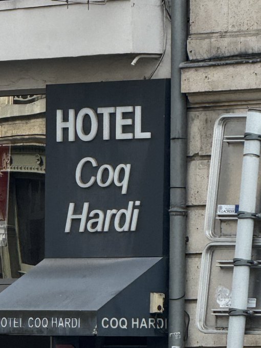 A black sign with white lettering reads "HOTEL Coq Hardi" mounted on a building, with the name repeated on the awning below. The name translates from French to "Proud Rooster Hotel," giving the awkward caption a fitting punchline.