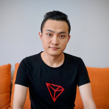 The image features a young man wearing a black shirt with a red triangle design on it. He is sitting on an orange couch against a white wall, posing for the camera with a serious expression. The background of the photo shows that he is indoors in a living room setting.