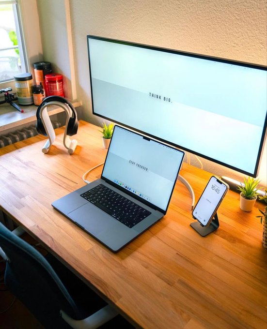 A wooden desk setup featuring a laptop and monitor both displaying 'THINK BIG.', alongside headphones on a stand, a phone showing 15:45, and small potted plants.