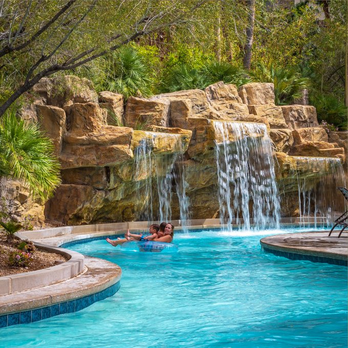 A couple relaxing on a tube float in a resort pool featuring a rock waterfall and tropical landscaping.