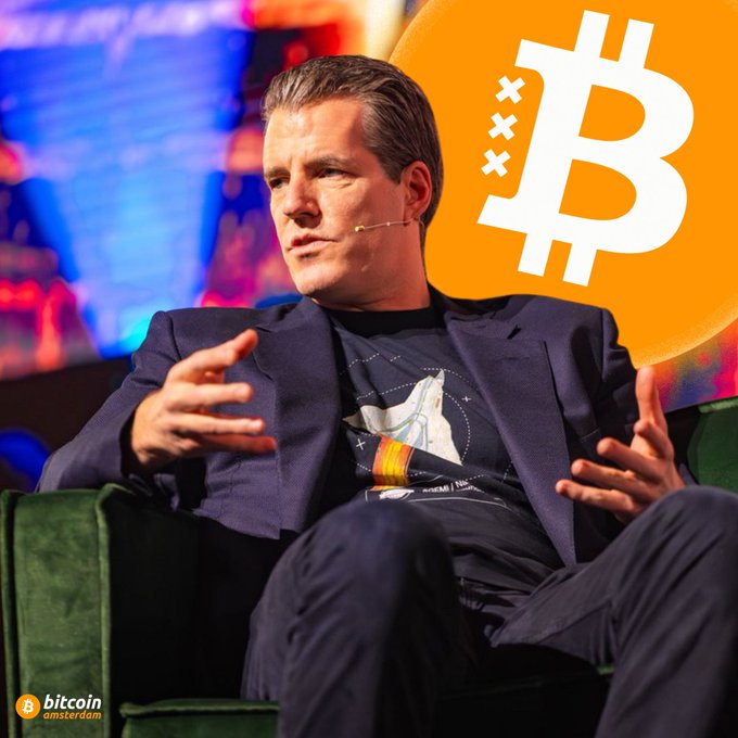 A man sits in a green chair on stage with a Bitcoin logo behind him, with a 'bitcoin amsterdam' watermark in the corner.