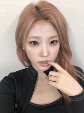 A close-up selfie of a young woman with long, coppery-red hair wearing a black top, posing with her finger near her lips.