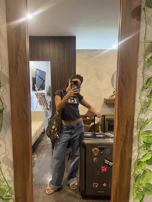 A woman poses for a mirror selfie in casual attire—a cropped "25" jersey tee, wide-leg jeans, and a monogram-patterned bag—while gripping a suitcase decorated with various travel stickers and a Bitcoin logo, hinting at her travel theme. The setting appears to be a modern lounge or hotel interior, reinforcing her teaser about heading to a Bitcoin-themed destination.