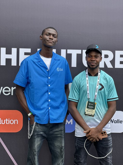 Two young men pose for a photo in front of a step-and-repeat banner; one wears a blue 'Sui' button-up shirt and the other wears a teal Converse jersey with a lanyard.