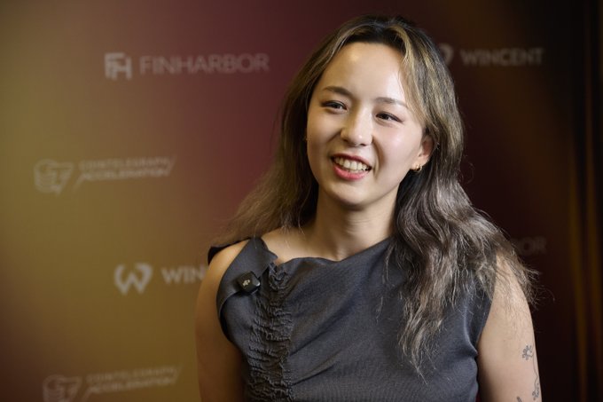 A young woman with long dark hair and a small tattoo on her arm smiles during an interview at a Finharbor event, wearing a dark sleeveless top with a microphone clipped to it. The backdrop features logos for "FINHARBOR" and "WINCENT," indicating a professional or business setting. Her appearance aligns with the post's mention of @sui414 from Pantera Capital, who discusses evaluating digital asset treasury (DAT) companies based on crypto holdings per share.
