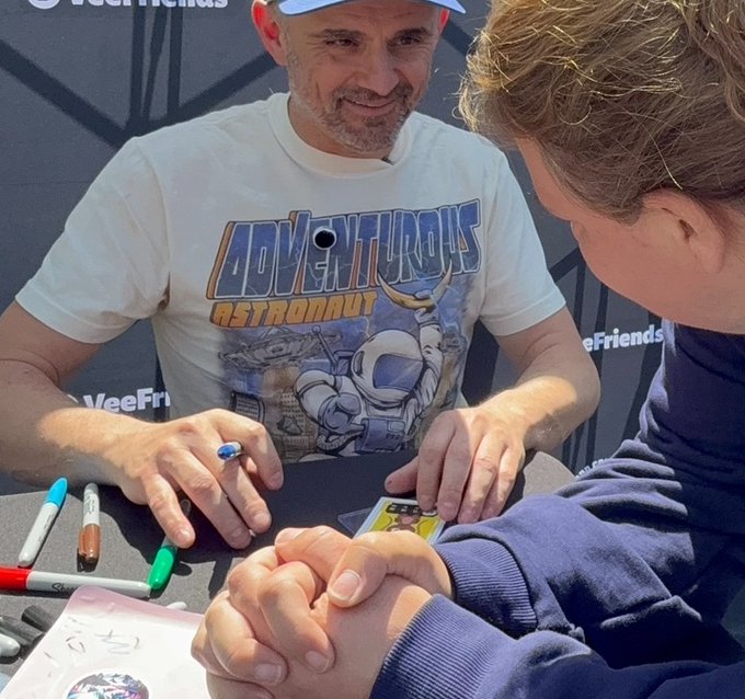 An older man with a gray beard wearing an 'Adventurous Astronaut' t-shirt smiles while sitting at a table signing autographs for a young fan.