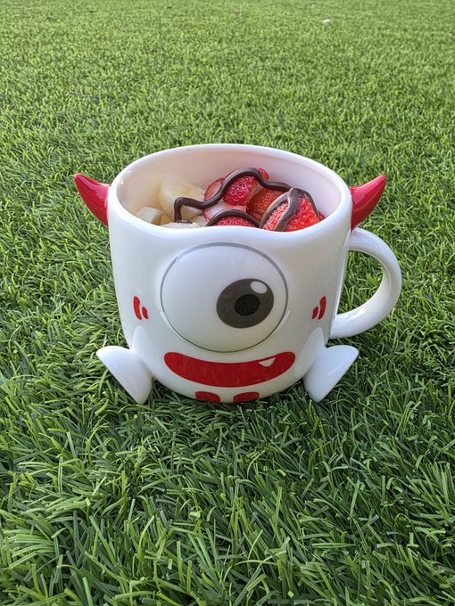 A novelty monster-shaped mug filled with cereal sits on artificial grass, featuring one large eye, red horns, and a toothy smile.