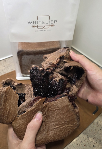 A pair of hands pulling apart a chocolate blowtorched bagel, revealing a dark, gooey filling, with a 'WHITELIER' branded bag visible in the background.