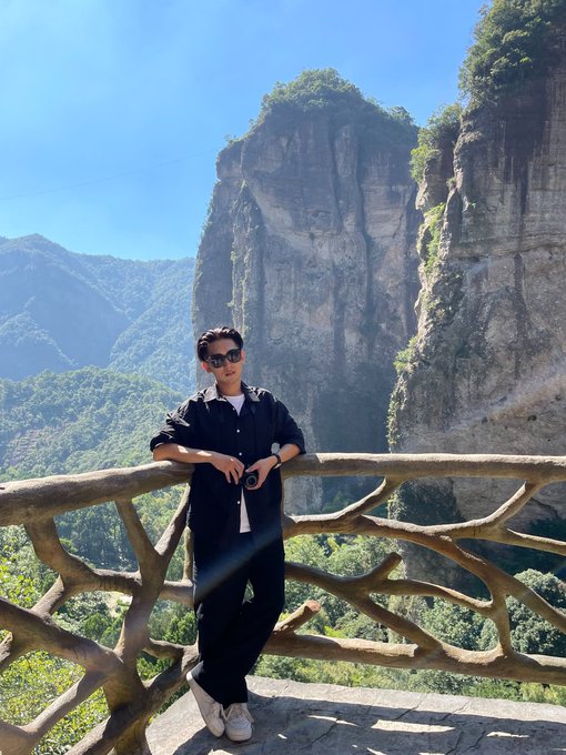A young man in sunglasses and dark casual wear poses confidently at a scenic mountain viewpoint, embodying the adventurous, ambitious spirit of a content creator carving out his own path.