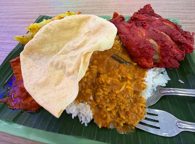 A plate of food served on a banana leaf, containing white rice covered with a yellow-orange curry, papadum, fried chicken, and tandoori chicken.