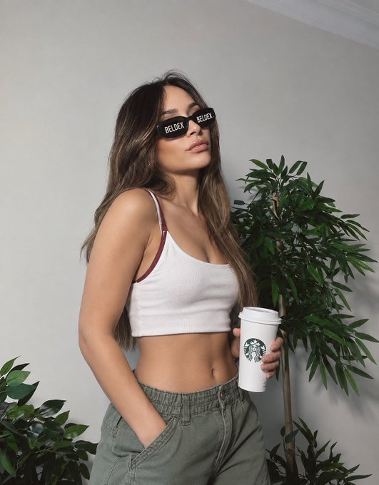 A young woman wearing sunglasses emblazoned with "BELDEX" holds a Starbucks coffee, modeling a casual white crop top and olive cargo pants beside a potted plant. The image complements the post by visually reinforcing the Beldex brand, tying the cryptocurrency's BNS (Beldex Name Service) feature to an everyday, approachable lifestyle aesthetic.