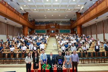 A wide shot of a large auditorium filled with a seated audience wearing masks, with several people posing in the front row above a 'bitkub' logo banner.