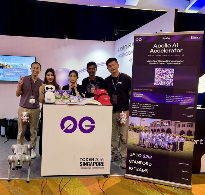 A group of five people pose behind a counter labeled '0G TOKEN2049 SINGAPORE 7-9 OCTOBER 2025 - MARINA BAY SANDS' next to a banner for the 'Apollo AI Accelerator' program.