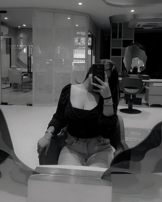 A black and white mirror selfie of a woman wearing a black top and jeans, sitting in a salon chair while holding her phone.